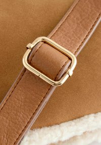 Cintura in finta pelle marrone con una fibbia quadrata dorata, superficie testurizzata, attaccata a un corpo in suede beige con foderatura bianca soffice.
