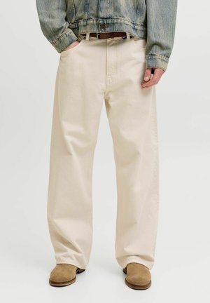 Osoba nosząca luźne kremowe spodnie, brązowy pasek, beżowe zamszowe buty oraz krótką niebieską kurtkę dżinsową z rękami w kieszeniach.