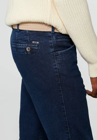 Donkerblauwe spijkerbroek met een slim fit, met een achterzak met een merklabel en een knoopdetail, gedragen met een beige geweven riem.