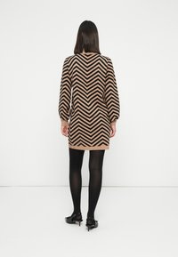 Kvinde iført en brun og sort zigzag-mønstret lang sweaterkjole, sorte strømpebukser og sorte kitten heel sko, stående opad en hvid væg.