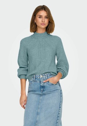 Turkizno pleteno pulover s rebrasto teksturo in visokim ovratnikom, v kombinaciji s svetlo modrim denim krilom. Obe oblačili imata priston oblik.