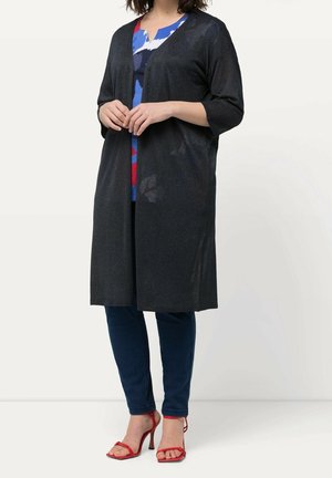 Mujer que lleva una chaqueta negra hasta la rodilla sobre una blusa colorida, vaqueros azul oscuro y sandalias rojas de tacón alto, de pie frente a un fondo liso.