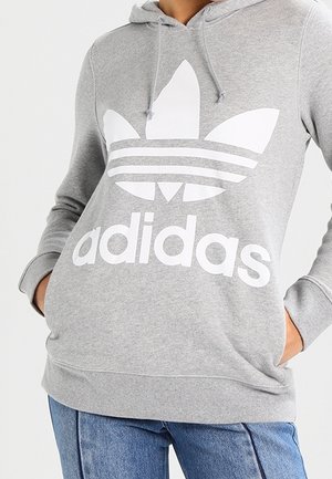 Persoon draagt een grijze Adidas hoodie met groot wit logo en kangoeroezak, gecombineerd met een blauwe spijkerbroek, handen in de zakken.