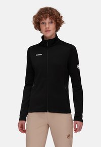 Giacca in pile nera con colletto alto, chiusura con zip, due tasche laterali e logo bianco. Texture liscia con un design aderente.