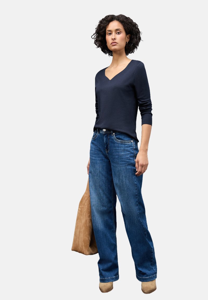 Top lungo a maniche lunghe blu navy con scollo a V, vestibilità rilassata, abbinato a jeans blu a gamba larga. La modella tiene una borsa tote in suede beige, in piedi su uno sfondo bianco.