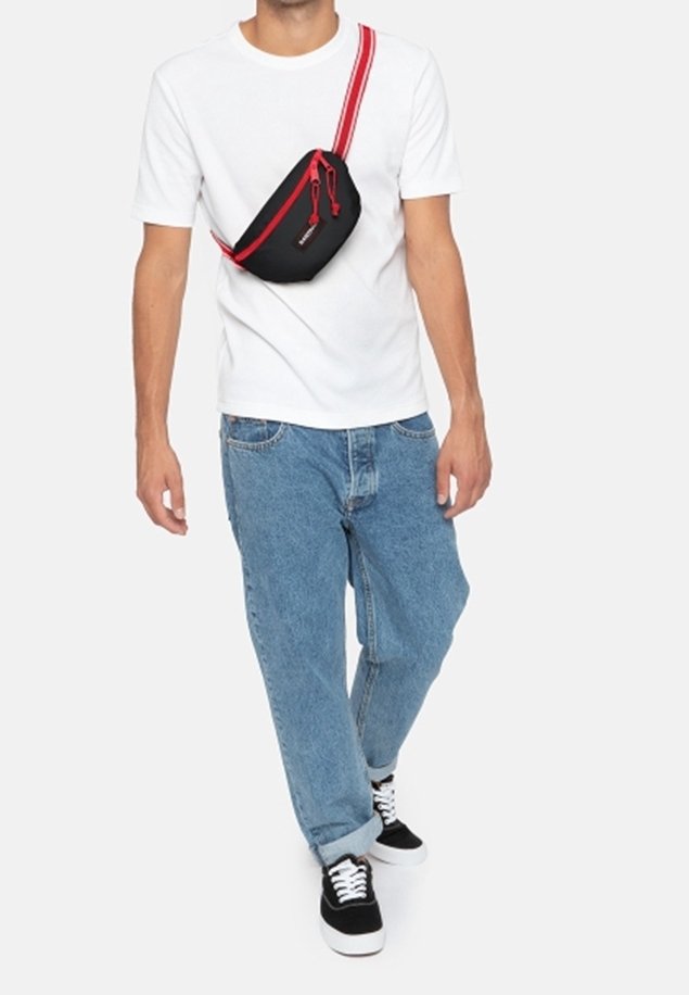 Black shoulder bag with a red strap and zipper, worn diagonally across a white t-shirt and blue jeans, featuring a casual design.