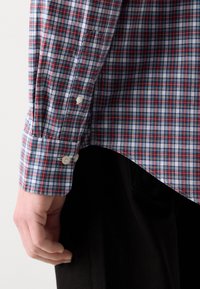 Gros plan d'une main reposant sur le côté portant une chemise à manches longues avec poignets boutonnés à motif à carreaux rouge, blanc et bleu et un pantalon noir.