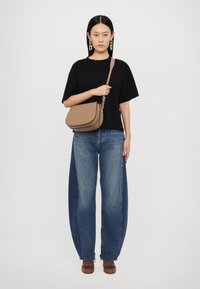 Borsa a tracolla marrone con una texture liscia, design curvo e dettagli in hardware dorato, abbinata a jeans blu oversized e una maglietta nera.
