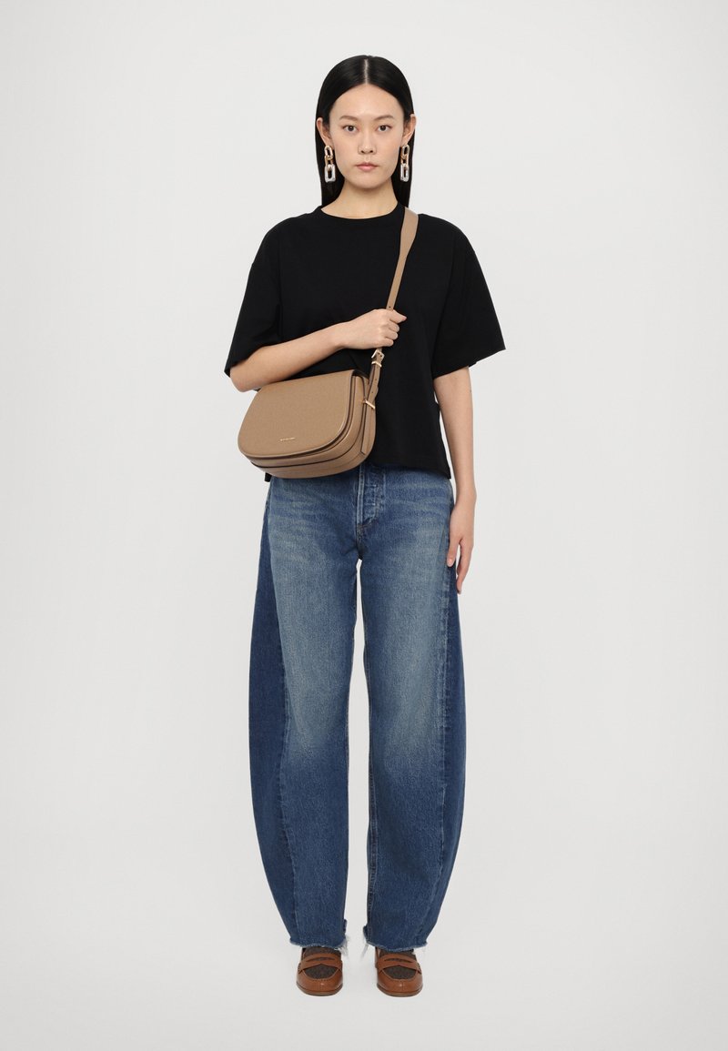 Borsa a tracolla marrone con una texture liscia, design curvo e dettagli in hardware dorato, abbinata a jeans blu oversized e una maglietta nera.