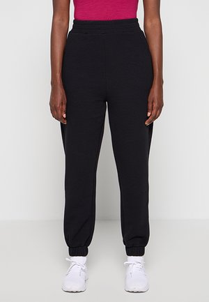 Osoba nosząca czarne teksturowane spodnie dresowe z elastycznymi mankietami, czerwoną bluzkę i białe buty sportowe, stojąca na jednolitym tle.