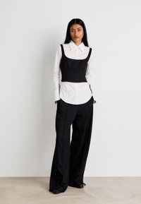 Top court noir texturé superposé à une chemise blanche à boutons, associé à un pantalon large taille haute noir avec des accents de boutons.