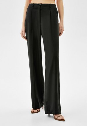 Femme portant un pantalon noir taille haute à jambes larges et des sandales à talons marron, debout devant un fond blanc uni.