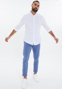 Camicia bianca con bottoni, pantaloni azzurri chiari, sneakers bianche. La camicia presenta un piccolo logo sul petto. Tessuto morbido, maniche arrotolate.