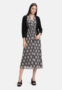 Robe maxi avec un motif paisley noir et crème, dotée d'une taille cintrée, d'un décolleté en V et d'un tissu léger et fluide. Accompagnée d'un cardigan noir.