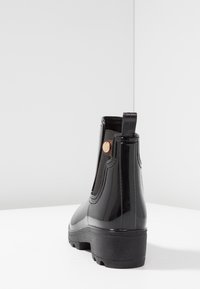 Musta kiiltävä nilkkurit joustavilla sivupaneeleilla, vetokahvalla ja teksturoidulla mustalla kumipohjalla. Koristeena pieni pronssinvärinen yksityiskohta aivan kantapään lähellä.