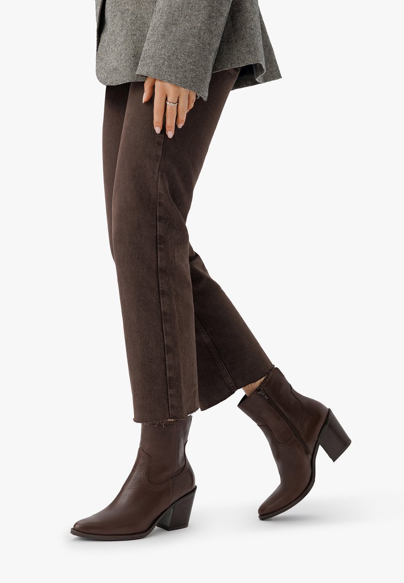 Bocanci de piele maro la gleznă, cu vârf ascuțit și toc îngrădit, având un finisaj texturat subtil și detalii cu fermoar lateral, purtați cu pantaloni maro închis.