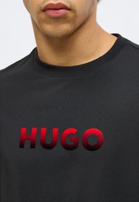 Čierne pletené tričko s výrazným červeným gradientovým logom "HUGO" s textúrovaným povrchom. Klasický okrúhly výstrih a krátke rukávy.