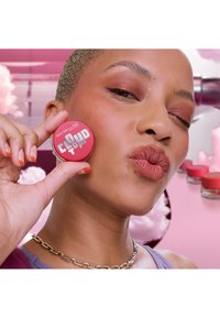 Personne aux cheveux courts tenant un produit pour les lèvres Maybelline Cloud Top rouge, portant un fard à paupières et un rouge à lèvres roses, sur un fond rose.