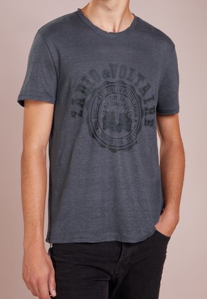 Homme portant un t-shirt Zara & Voltaire gris foncé à manches courtes avec un col rond et un logo avant délavé, pantalon sombre, main dans la poche.
