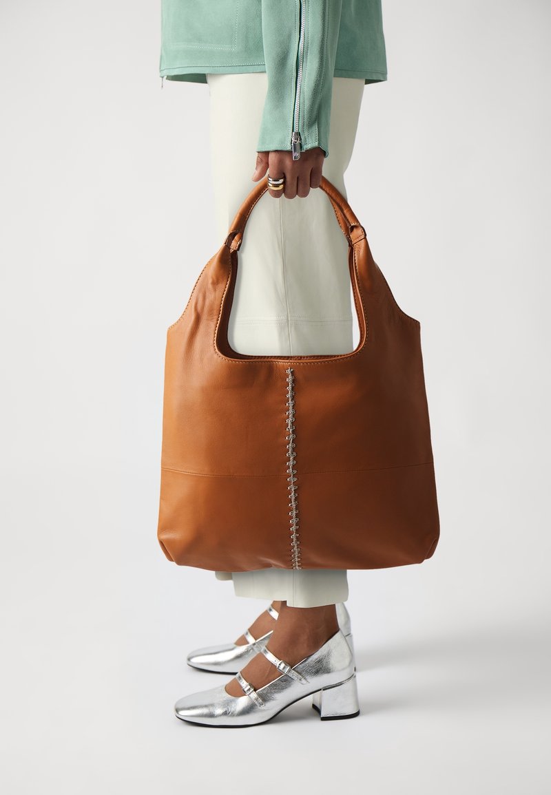 Personne tenant un grand sac à main en cuir marron avec une couture centrale, portant un pantalon blanc, une veste verte zippée et des chaussures Mary Jane argentées.
