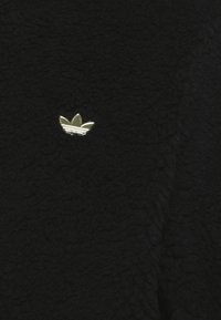 Svart fleece-tyg med ett litet guldemblem med tre blad och ränder i mitten, som visar en texturerad yta.