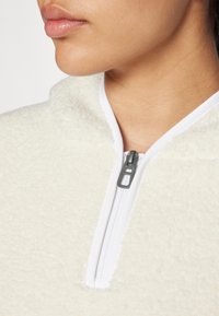 Pull en polaire à demi-zip de couleur crème avec une surface texturée et duveteuse et une fermeture éclair blanche. Vue rapprochée des détails du col et de la fermeture éclair.