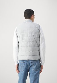 Ljust grå vadderad väst med horisontella sömmar, rundad krage och logotypdetalj. Bärs över en vit långärmad skjorta och blå jeans.