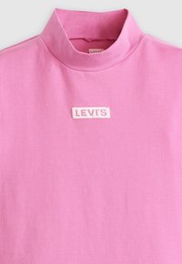 Vaaleanpunainen Levi's-t-paita korkealla kauluksella, keskellä valkoinen kudottu logo-patch. Silkkinen puuvillakangas, minimalistinen muotoilu.