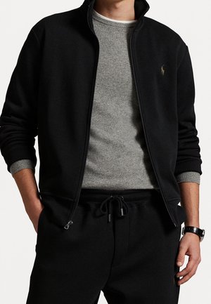 Mann trägt schwarze Reißverschlussjacke über grauem Pullover und schwarze Jogginghose mit Kordelzug, Armbanduhr am linken Handgelenk, Hände in den Taschen.