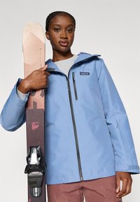 Αδιάβροχο μπουφάν σκι σε ανοιχτό μπλε με φερμουάρ μπροστά, σε συνδυασμό με καφέ παντελόνι σκι, κρατώντας ένα ροζ σκι με μαύρους δεσίματα.