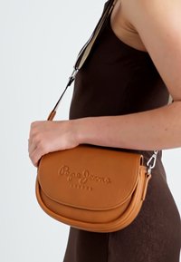 Hnědá kožená crossbody taška s zakřiveným tvarem, embosovaným logem a odnímatelným popruhem. Povrch je hladký s minimálními kovovými prvky.