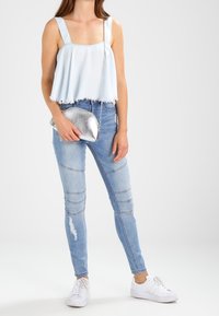 Ljusblå, kort, ärmlös topp med fransar i nederkant; kombinerad med figurnära, slitna jeans och vita sneakers; silverfärgad clutch som accessoar.