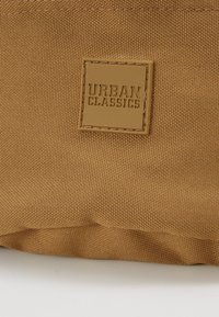 Sacchetto di stoffa marrone con superficie testurizzata e una toppa in pelle quadrata con "URBAN CLASSICS" impresso in caratteri in grassetto.