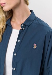 Mörkblå skjorta med knappar i mjukt, lätt tyg med en diskret broderad logotyp på vänster bröst och klara plastknappar.