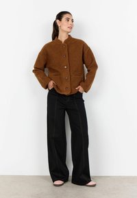 Hnedá chlpatá bunda s vysokým golierom, predným zapínaním na gombíky a dvoma vreckami, v kombinácii s čiernymi širokými nohavicami a ružovými balerínami.