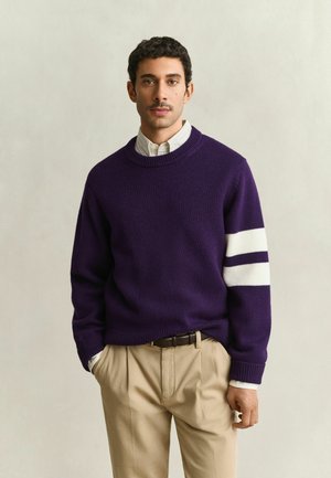 Uomo che indossa un maglione viola con due strisce bianche su una manica, una camicia bianca con colletto e pantaloni beige, con le mani in tasca.