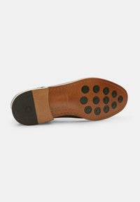Suola di scarpa in cuoio marrone con tappi di trazione neri in gomma rotondi, sezioni lisce e testurizzate, e un dettaglio di bordo grigio. Taglia 42.