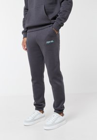 Uomo che indossa pantaloni da jogging grigio scuro con il logo "CLSR LDN Design Atelier" sulla coscia e scarpe da ginnastica bianche, in piedi con una mano in tasca.