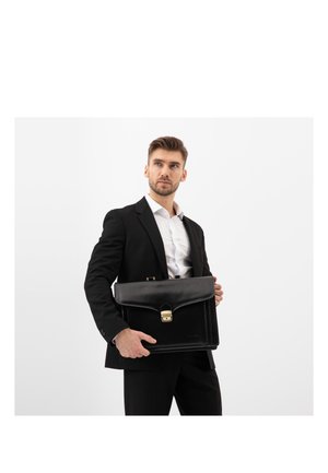 Uomo in abito nero e camicia bianca che tiene una valigetta in pelle nera chiusa con fibbia dorata, che guarda leggermente di lato su sfondo bianco.