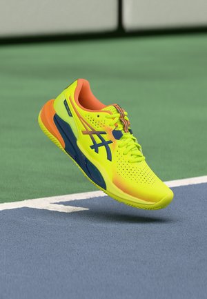 Dzeltens un oranžs sporta apavu pāris ar melnu logotipu peld virs zilās un zaļās tenisa korta virsmas netālu no baltās robežas līnijas.