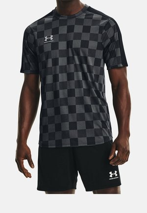 Mężczyzna ubrany w czarno-szarą, kraciastą koszulę z krótkim rękawem z logo Under Armour oraz jednolite czarne szorty z logo Under Armour.