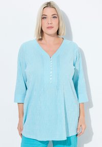 Lichtblauwe tuniek met verticale witte strepen, V-hals, driekwart mouwen en decoratieve knopenrij. Zachte, gestructureerde stof.