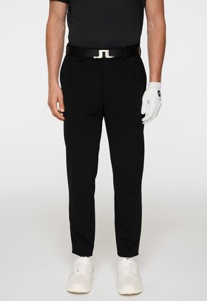 Zwarte golfbroeken met een getailleerde pasvorm, voorzien van een zwarte riem met een zilveren gesp, gecombineerd met een witte handschoen en witte sneakers.