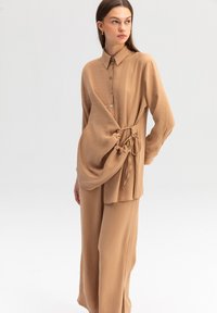 Blusa a maniche lunghe, marrone chiaro, con colletto, bottoni sul davanti e dettaglio laterale annodato, realizzata in un tessuto morbido e fluido.
