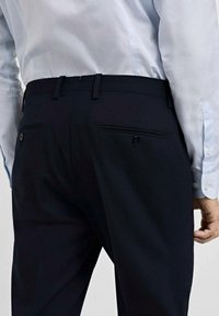 Marineblaue formelle Hose mit zwei Gesäßtaschen, die jeweils mit einem Knopf gesichert sind. Kombiniert mit einem hellblauen Hemd, das über Manschettenknöpfe verfügt.