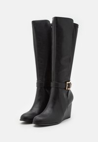 Une paire de bottes noires montantes à talons compensés avec fermeture éclair latérale et sangle décorative avec boucles dorées sur fond clair.