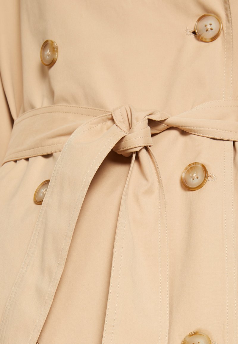 Trench beige con cinturón atado en la cintura, que presenta dos botones grandes y transparentes, y un tejido suave y texturizado.