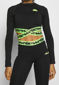Svart långärmad crop top med blixtlås, med reflekterande neongrön branding och detaljer. Stretchigt material, åtsittande design.