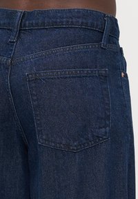 Jeans de mezclilla azul oscuro con una superficie texturizada, que cuentan con un bolsillo trasero y detalles de costura sutiles. La cinturilla es de altura estándar.