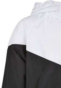 Veste coupe-vent blanche et noire montrant le haut du dos et la capuche avec un design à découpe diagonale des couleurs.